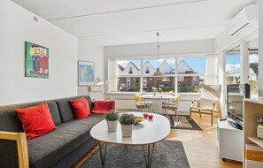 Una sala de estar moderna con un sofá gris, cojines rojos, una mesa redonda de centro con plantas, una zona de comedor con una mesa y sillas blancas, y ventanas amplias que ofrecen vistas a casas.