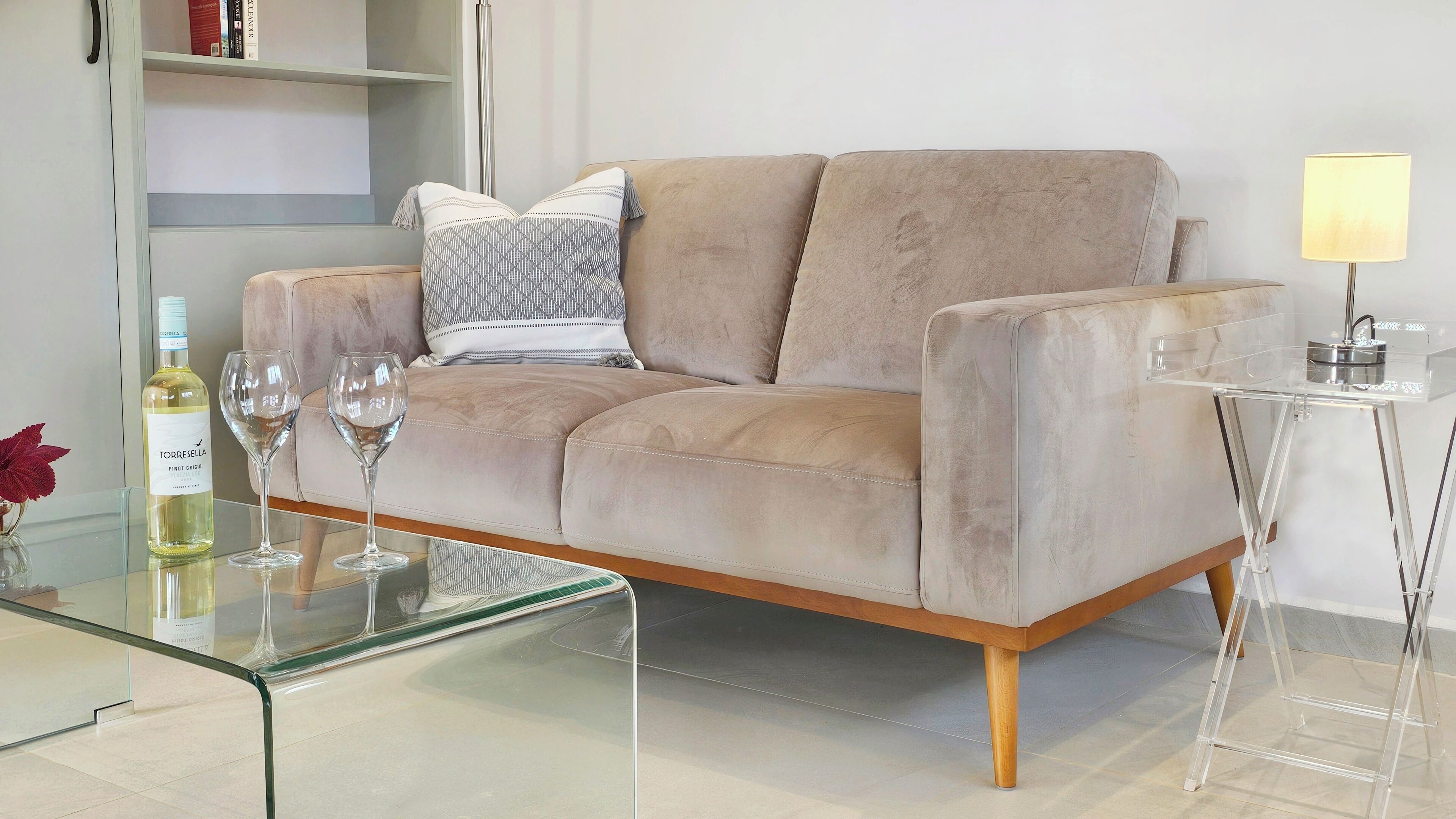 A beige sofa with a glass coffee table, a clear side table with a lamp, and a wine bottle and glass on the table.