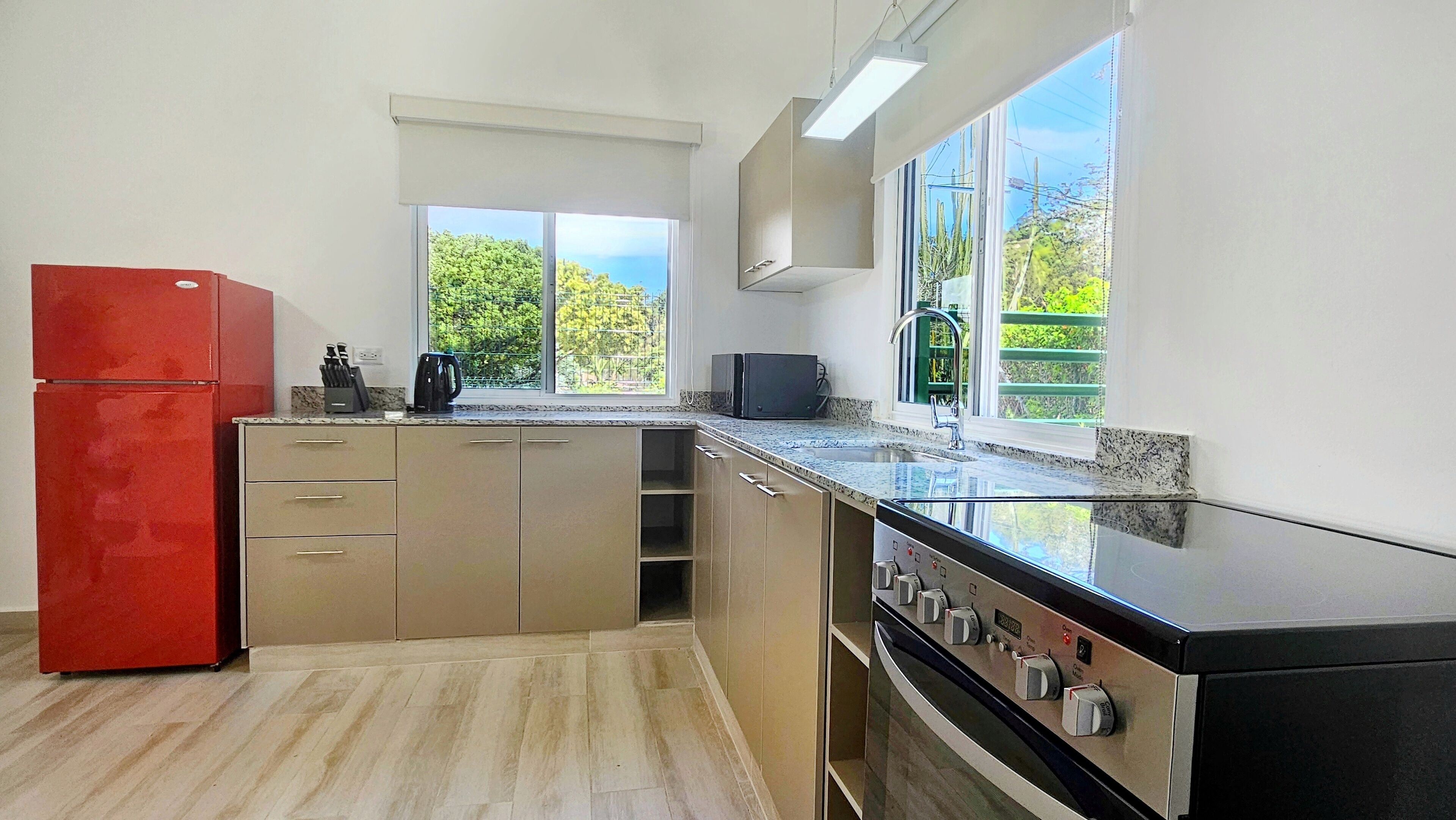 A modern kitchen with a red refrigerator, a built-in oven, and a window with a view of greenery.