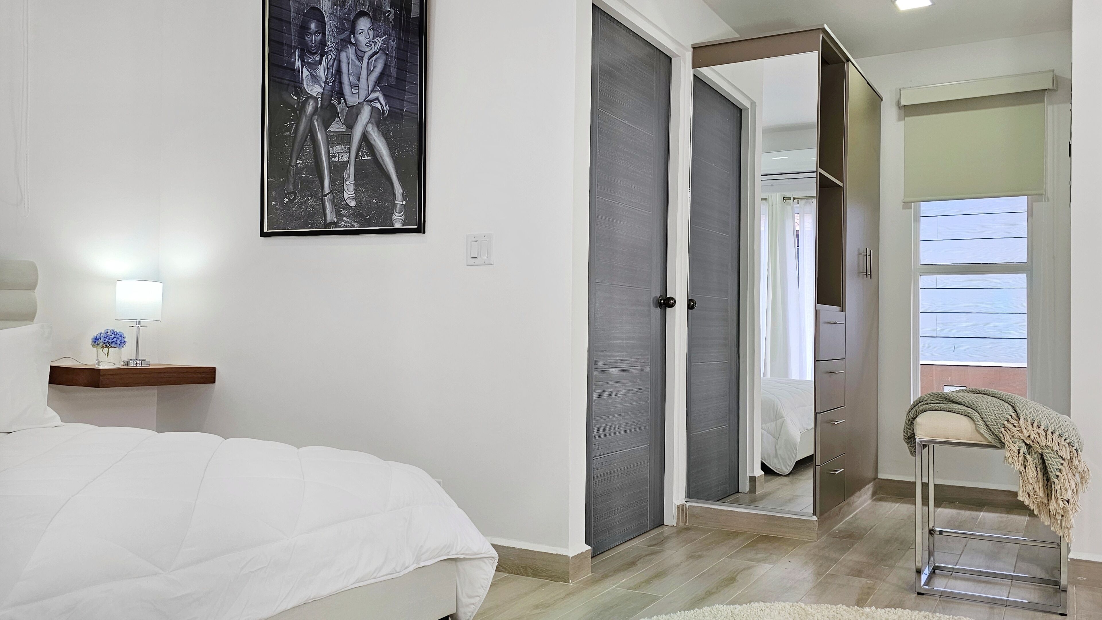 A bedroom with a bed, a bedside table with a lamp and flowers, a wardrobe, and a mirror.
