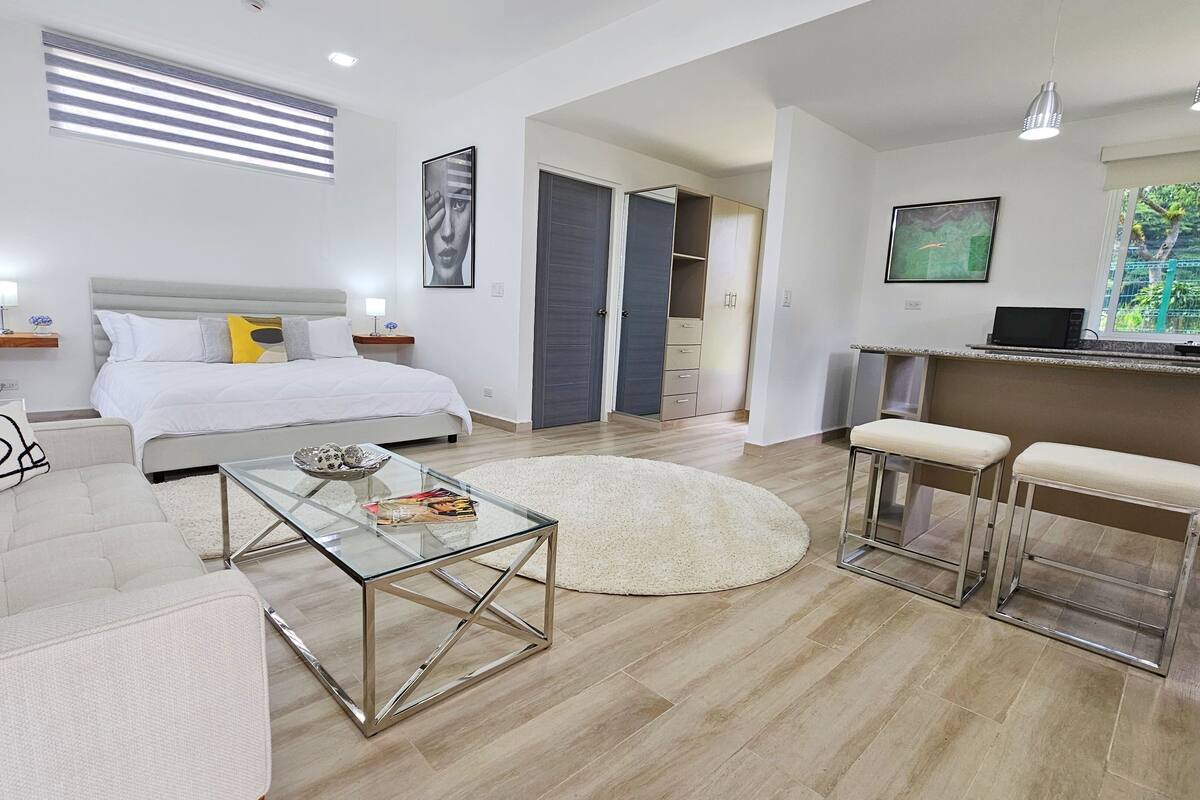 A modern hotel room with a bed, a glass coffee table, a sofa, and a kitchen area with a bar.