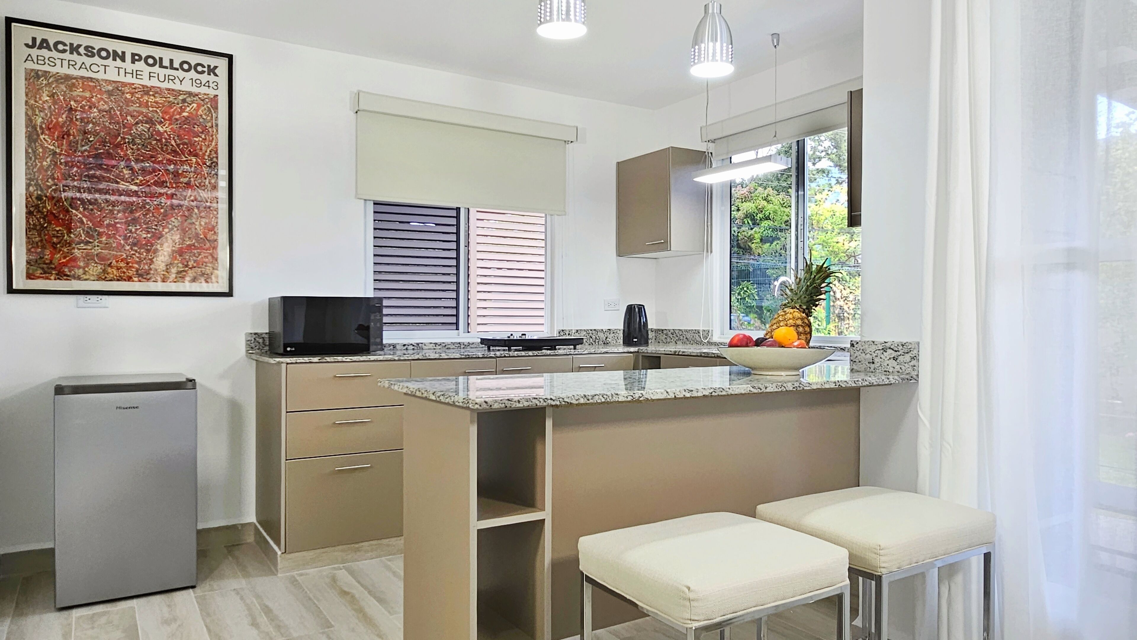A modern kitchen with a microwave, a coffee maker, and a fruit bowl. There is a small refrigerator, a window with blinds, and a chair.