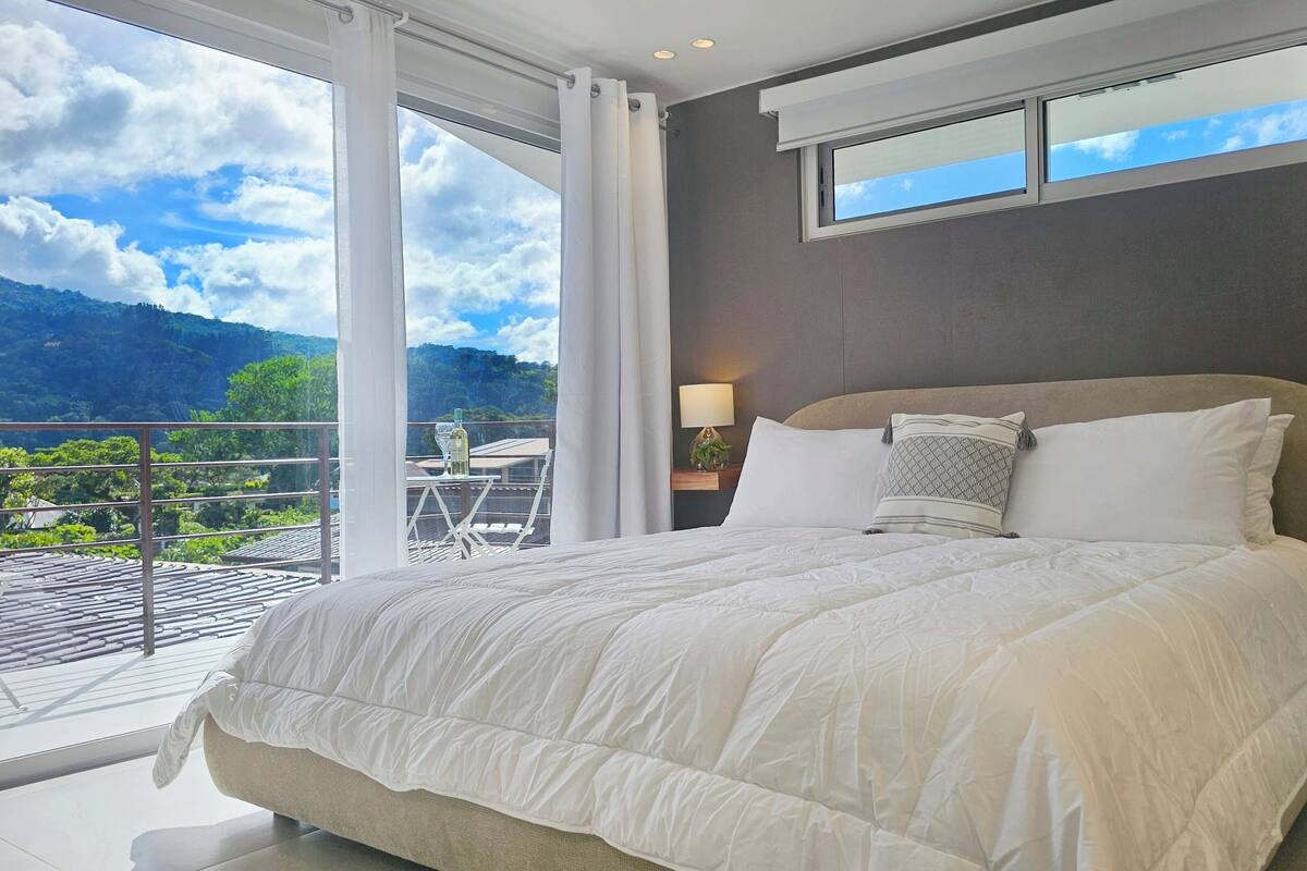 A bedroom with a large bed, a nightstand with a lamp, and a view of a balcony with a table and chairs.