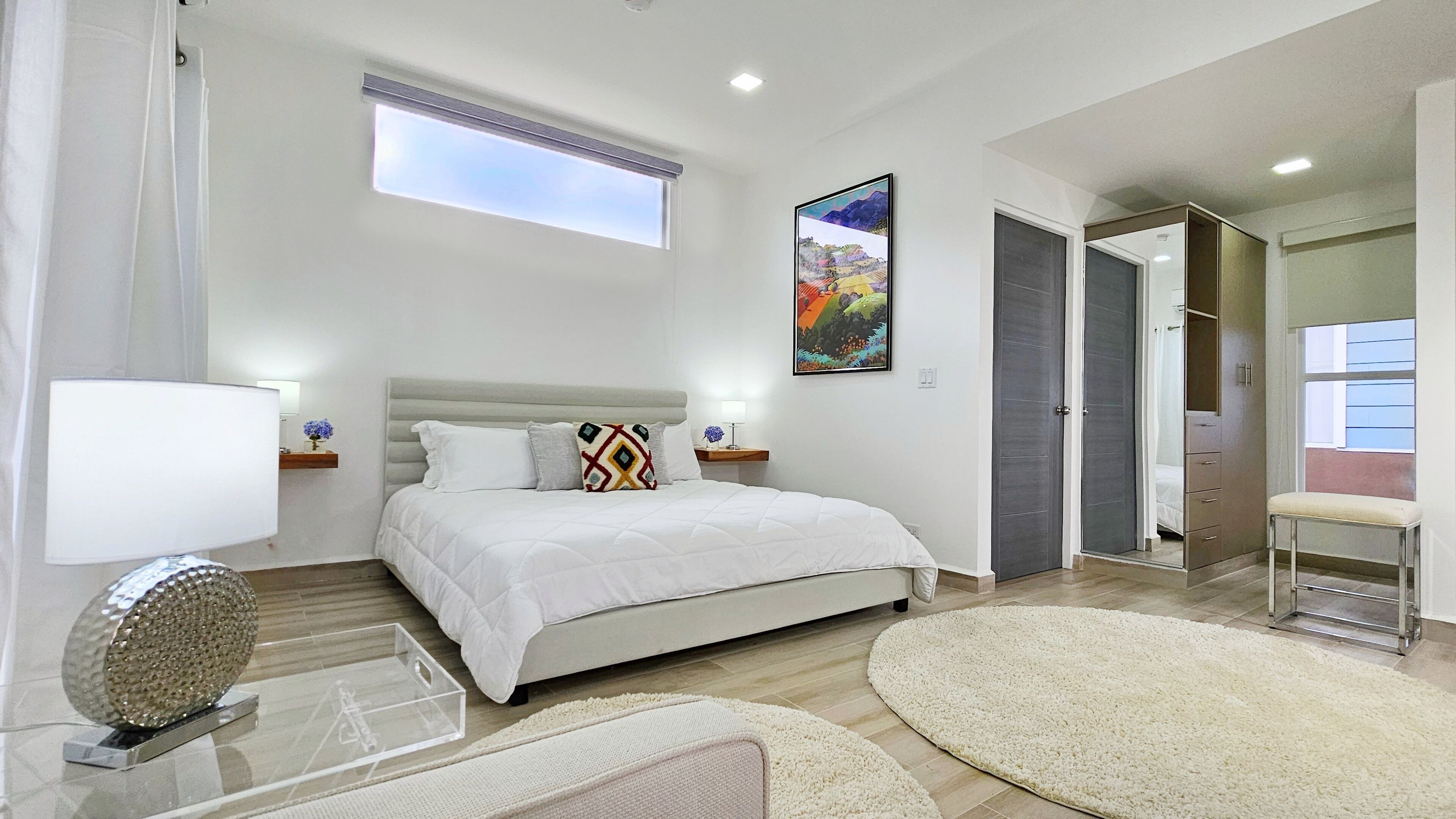 A modern bedroom with a large bed, a window, a painting, and a wardrobe.
