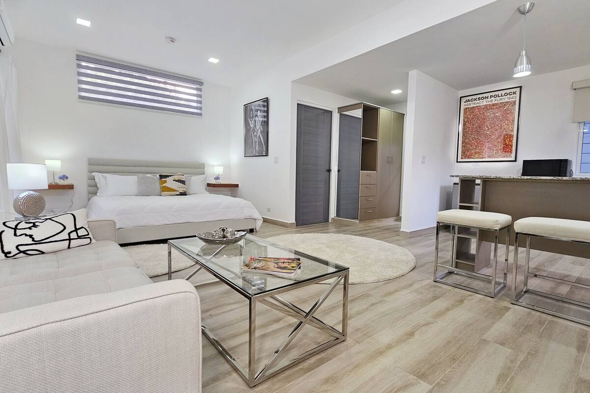 A modern, well-lit living room with a large bed, a glass coffee table, and a kitchen area with bar stools.