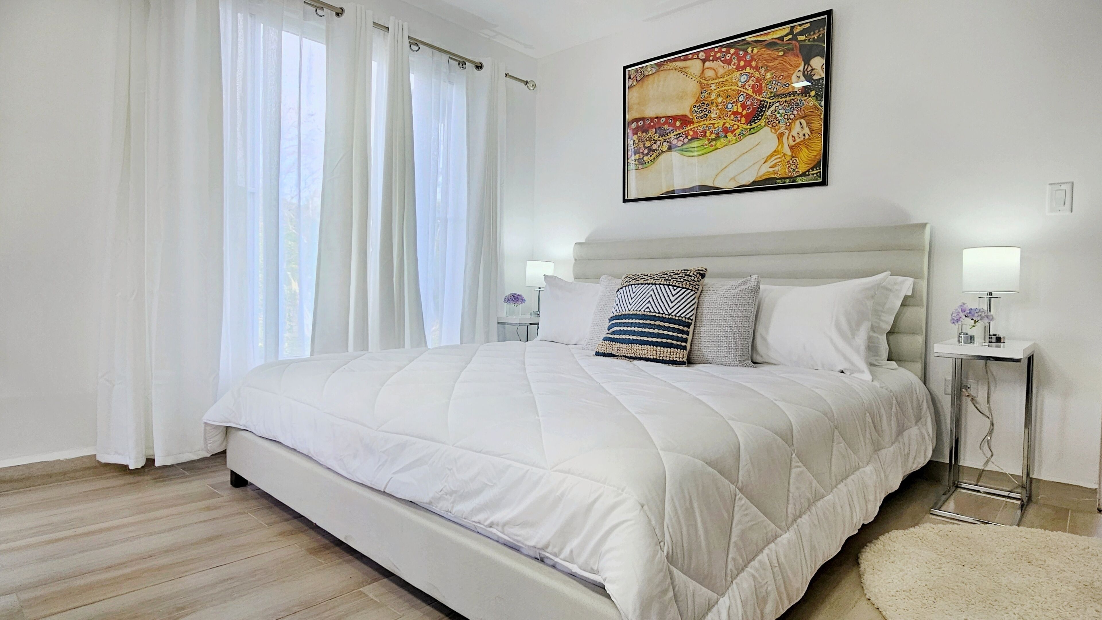 Un dormitorio moderno con una cama grande, ropa de cama blanca, cabecera, mesitas de noche con lámparas y un cuadro en la pared.