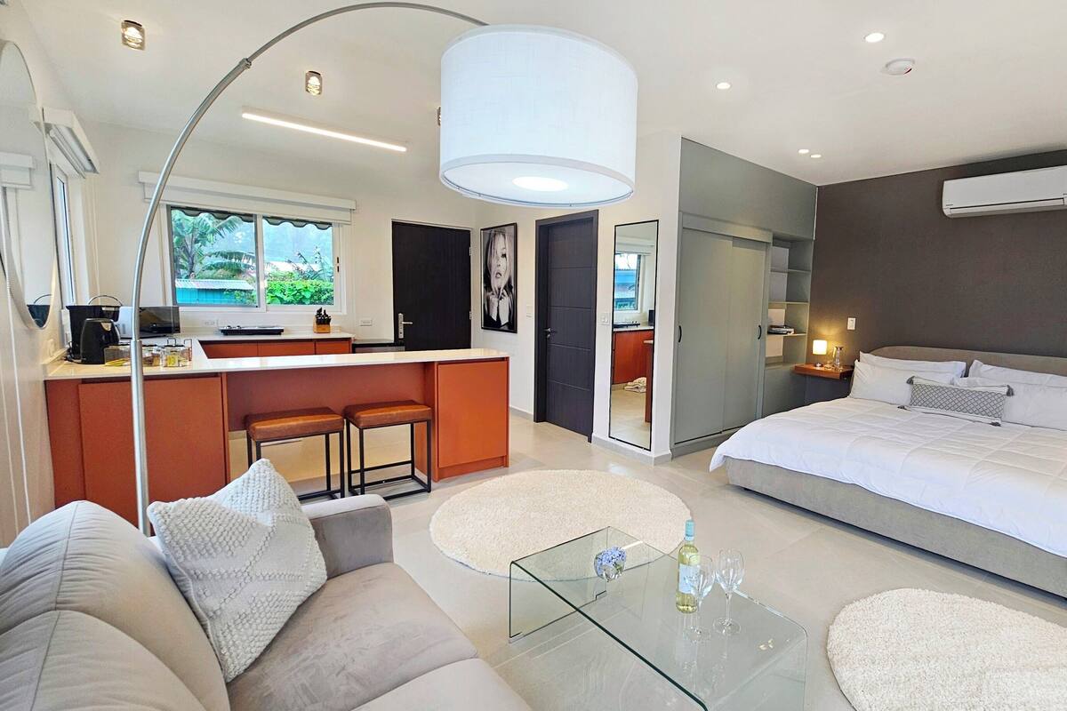 A modern hotel room with a large bed, a glass coffee table, a sofa, a kitchen area with a bar, and a view of the outdoors.
