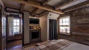 A log cabin-style bedroom with a fireplace, a bed, a TV mounted on the wall, and a window with curtains. - Welcome to Brooks Run - A 1920s Cabin in Beautiful Brown County, Indiana (Nashville)
