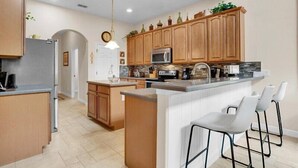 A modern kitchen with wooden cabinets, a central island with bar stools, a microwave, and a sink. - Magical Memories 5 BR Spacious Pool/Spa Home (Kissimmee)