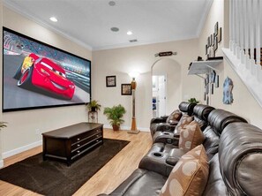 A living room with a large flat-screen TV, a dark brown entertainment center, a leather sofa with patterned cushions, a tall floor lamp, and a staircase with a white railing.