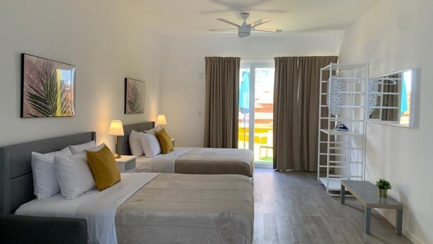Habitación de hotel con dos camas, ventilador de techo, balcón con cortinas y una estantería blanca.