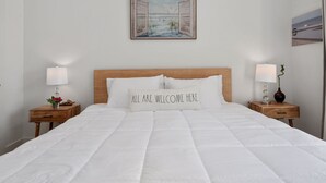 Una cama bien hecha con edredón blanco y almohadas, dos mesitas de noche con lámparas, un cabecero de madera y un cuadro enmarcado sobre la cama.