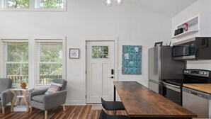 Una cocina moderna con una mesa y sillas de madera, un refrigerador, microondas y horno. Hay ventanas con persianas, una puerta y un estante en la pared con objetos.