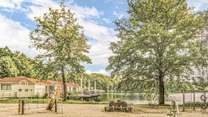 Un parque junto al lago con mesas de picnic, un área de juegos y un pequeño edificio con techo rojo.