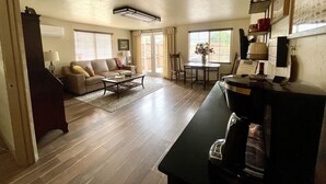Un'ampia zona giorno con divano, tavolino da caffè e tavolo da pranzo. C'è un angolo cottura con frigorifero e forno a microonde. La stanza presenta pavimenti in legno e una ventola a soffitto.