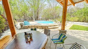 An outdoor patio with a wooden table and striped chairs, a stone wall, and a swimming pool in the background. - Beachfront Villa – Just Steps from the Sand, Your Ocean Escape Awaits (Guanacaste Province)