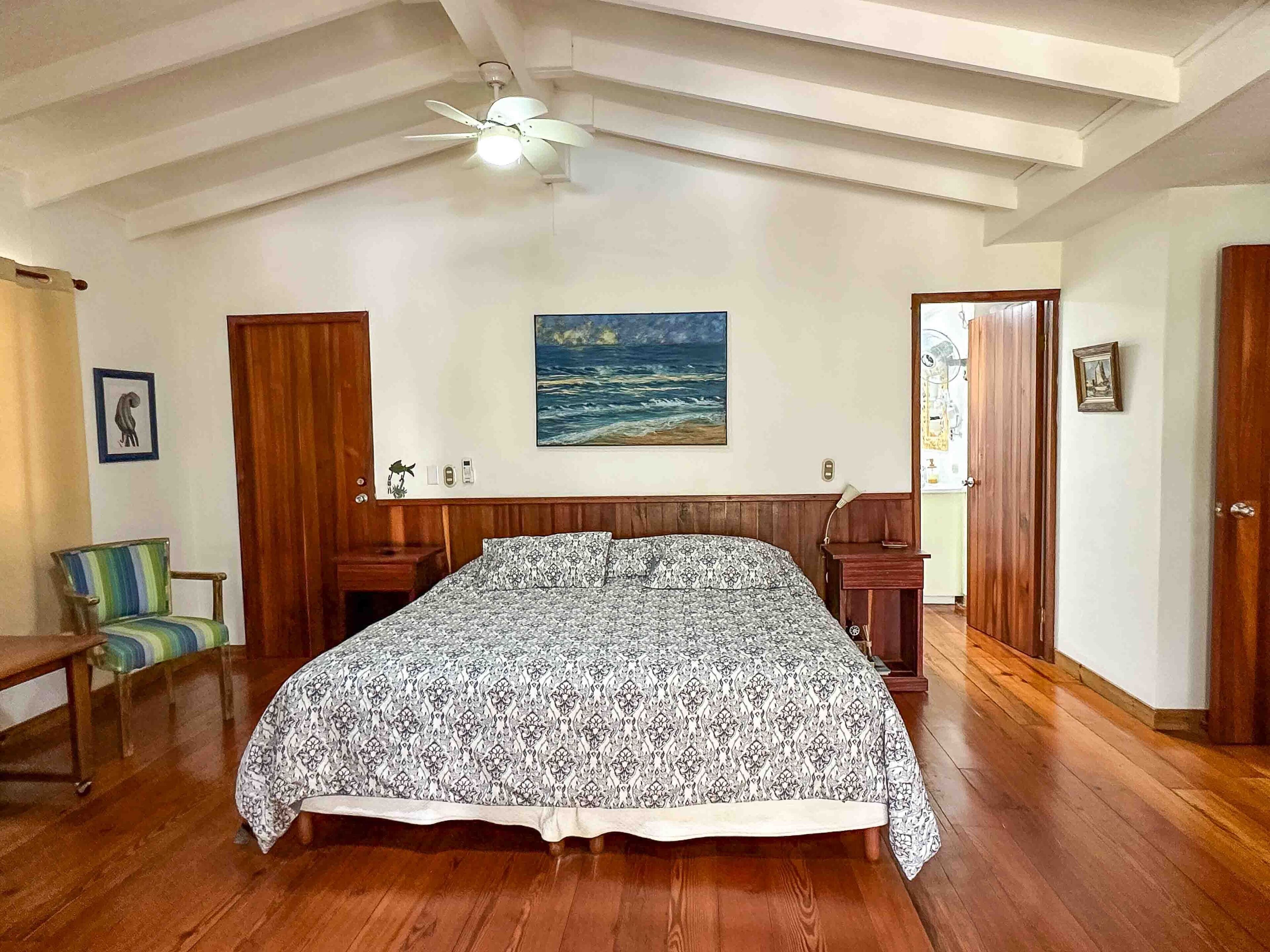 Une chambre avec un lit en bois, un ventilateur de plafond, un tableau représentant l’océan et des portes en bois.
