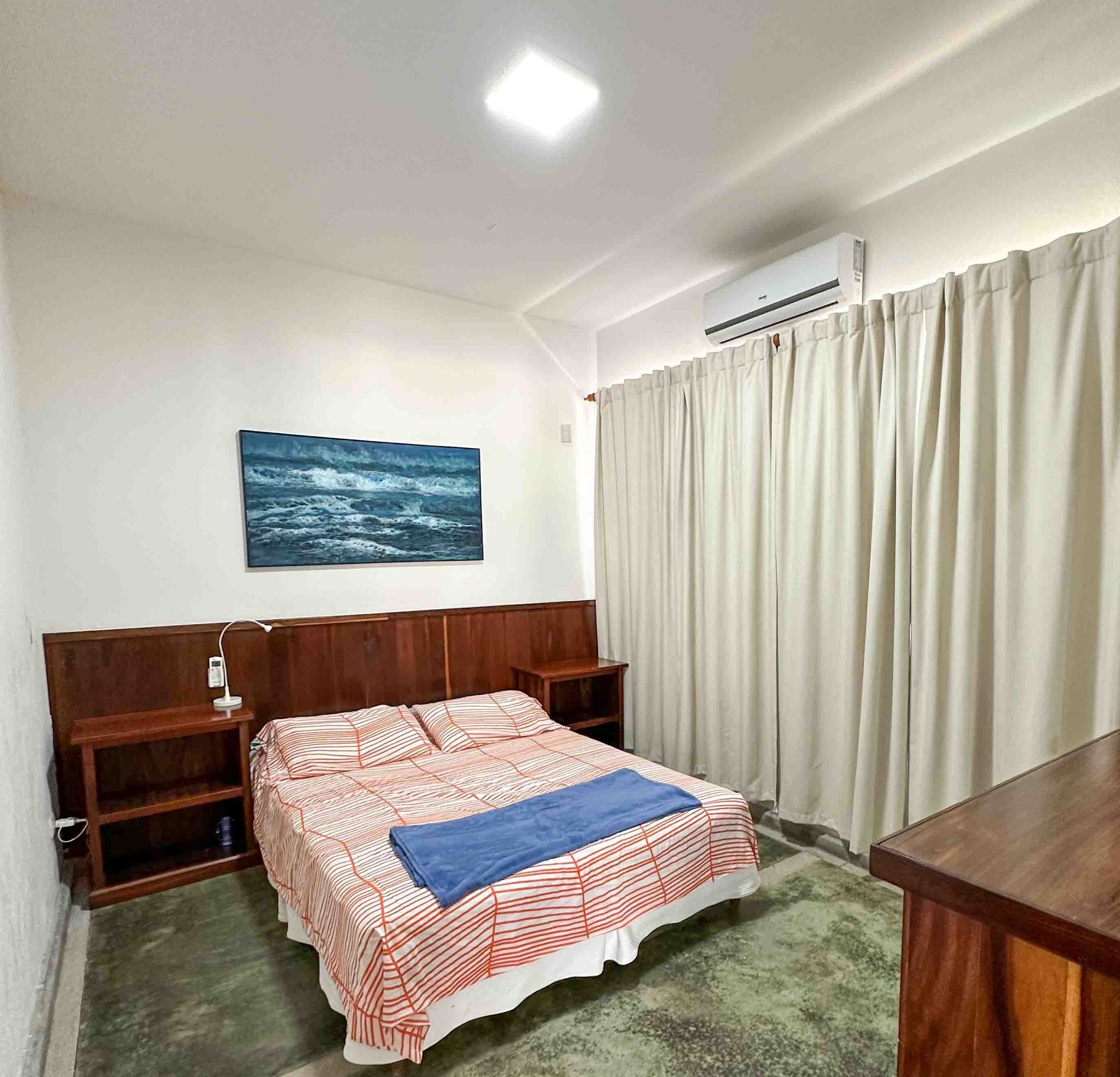 Un dormitorio con cama, escritorio, silla, armario, un cuadro y aire acondicionado.