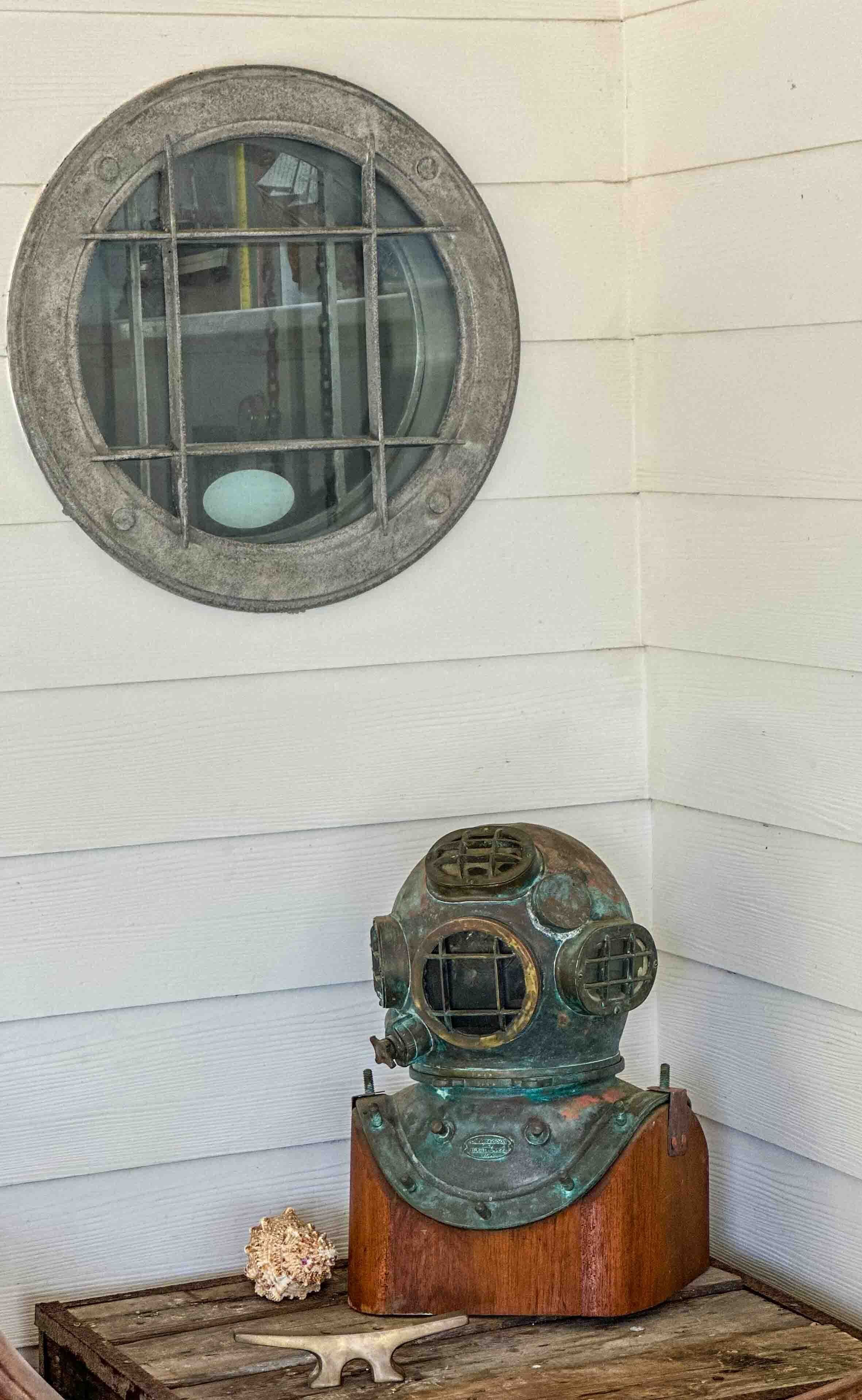 A vintage diving helmet on a wooden stand against a white wall with a round window above it.