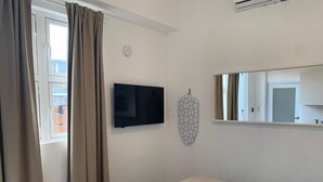 A hotel room with a bed, a TV mounted on the wall, a window with curtains, and a mirror. - Villa acogedora dentro del hospital Auxilio Mutuo 106 (San Juan)