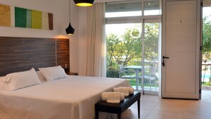 Une chambre d’hôtel avec un grand lit, un banc avec des serviettes, une tête de lit en bois, un luminaire au plafond et une porte donnant sur un balcon avec vue sur les arbres et une piscine.