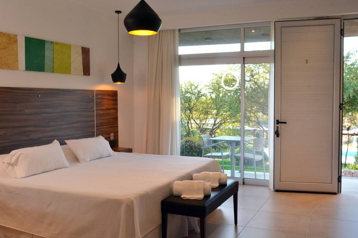 Une chambre d’hôtel avec un grand lit, un banc avec des serviettes, une tête de lit en bois, un luminaire au plafond et une porte donnant sur un balcon avec vue sur les arbres et une piscine.