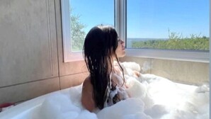 Une personne dans une baignoire remplie de mousse, avec vue sur un ciel bleu dégagé et une végétation luxuriante à l’extérieur.