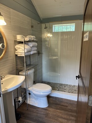 Ein Badezimmer mit Toilette, Waschbecken und Dusche. Handtücher liegen auf einem Ablagefach, und ein Schild weist auf einen Stromerraum hin.