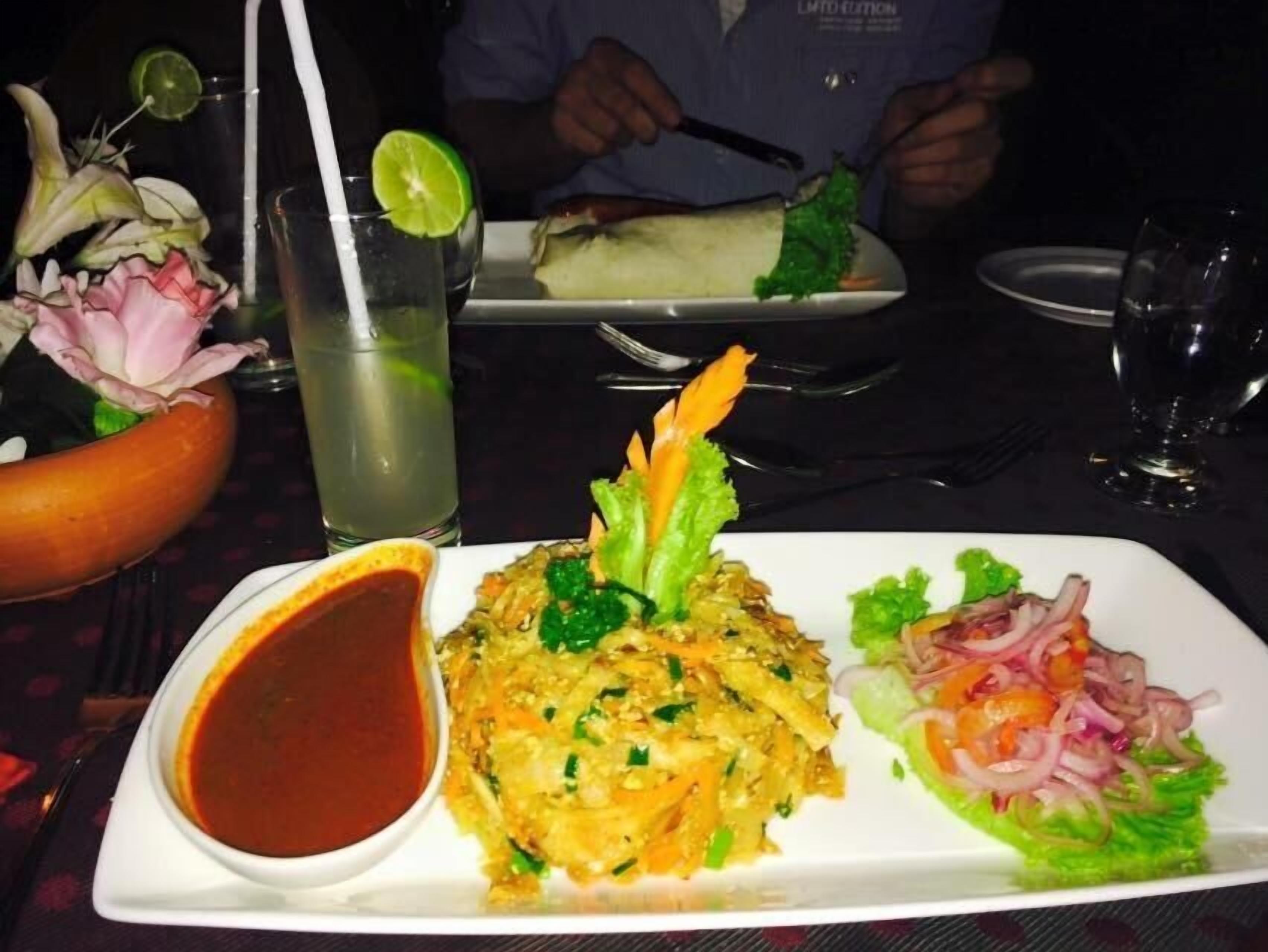 A plate of food with a side of sauce, a glass of drink, and a decorative flower arrangement.