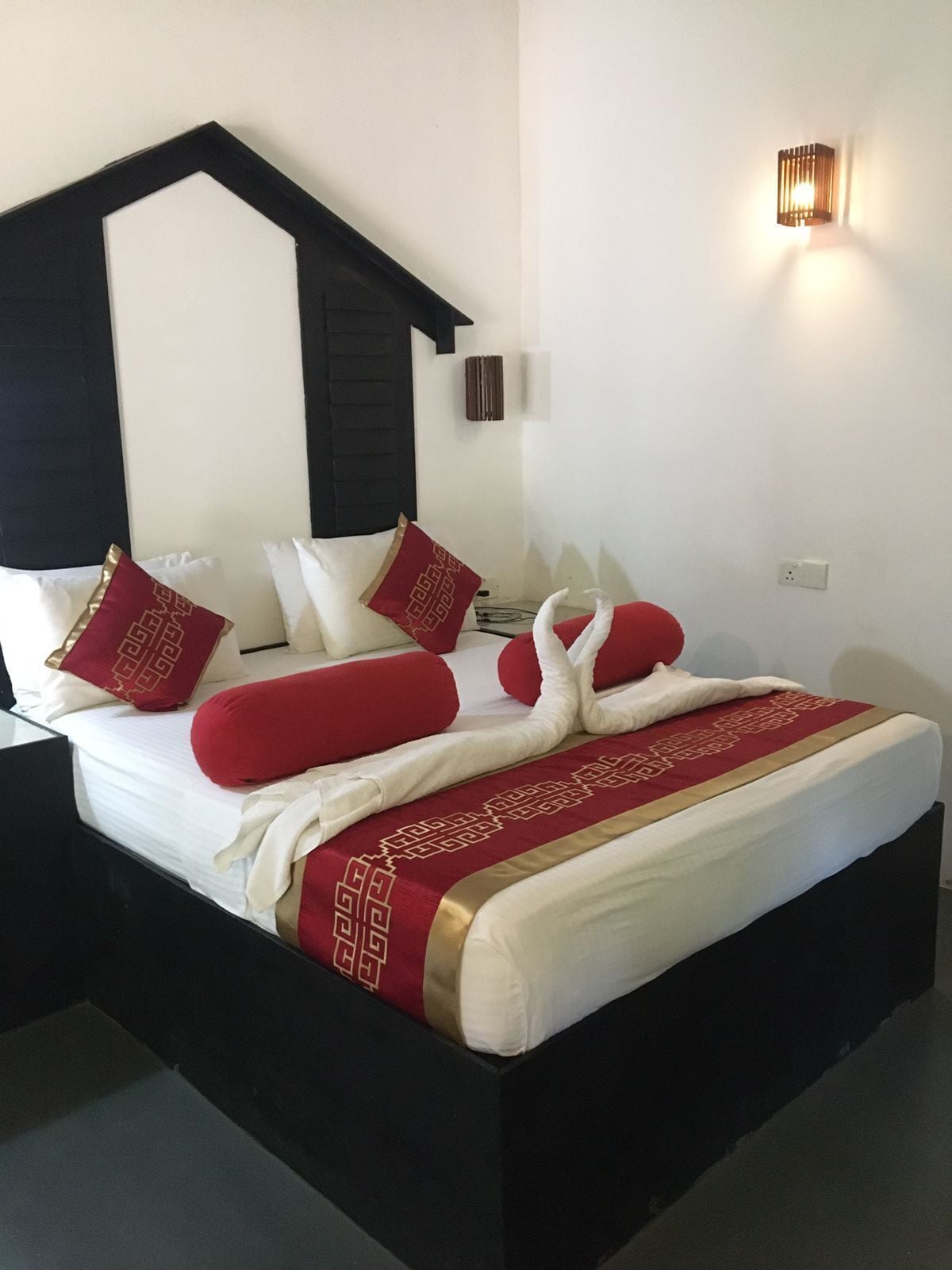 Una cama con estructura negra, ropa de cama blanca y almohadas rojas. Arriba, una lámpara fijada a la pared.