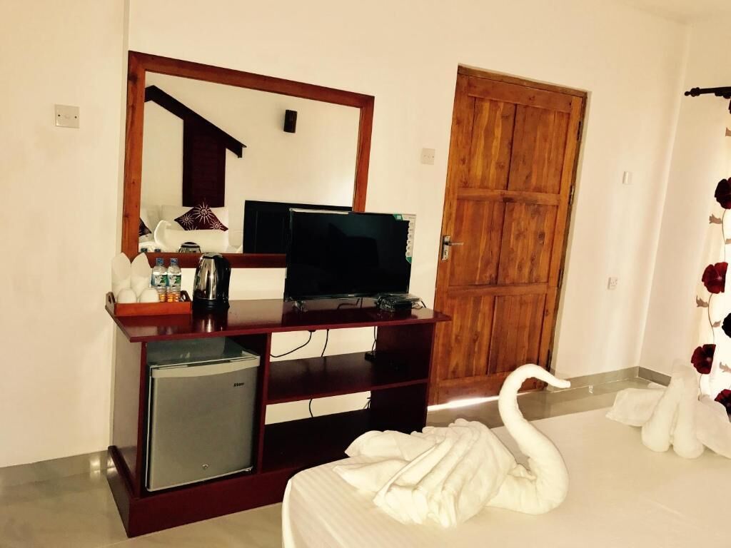 Habitación de hotel con cama, televisor sobre un atril de madera, refrigerador pequeño, puerta de madera y decoración de cisne en la cama.