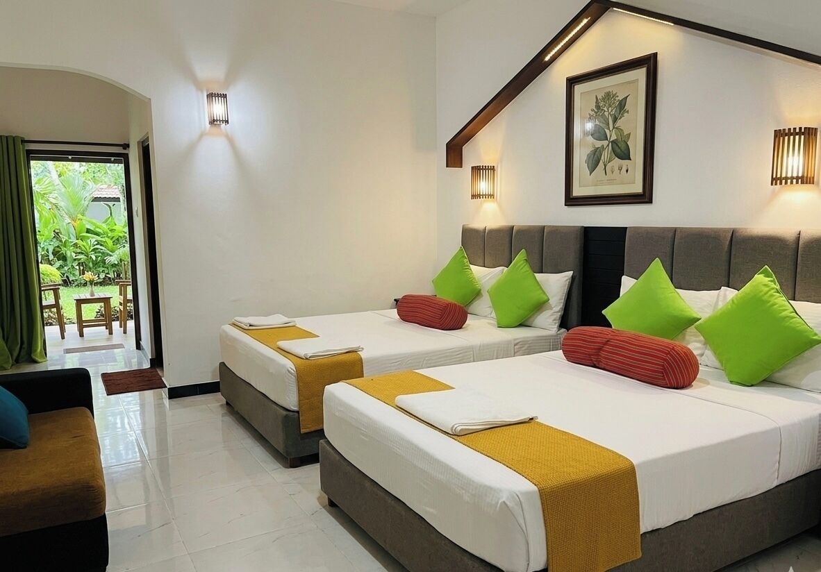A hotel room with two beds, a sofa, a dining table, and a balcony with a view of greenery.