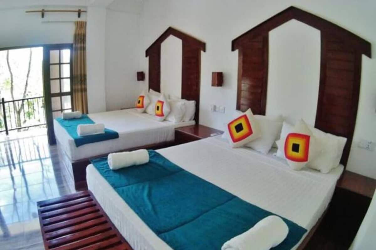 A hotel room with two beds, each with a wooden headboard, white bedding, and teal bedspreads. There is a wooden bench at the foot of the beds, and a window with a view of the outdoors.