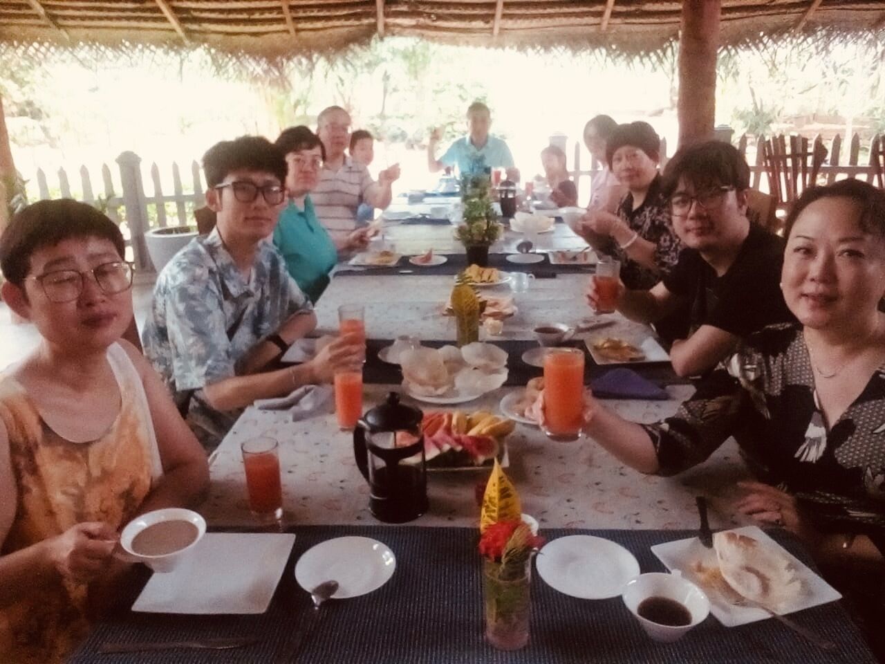 A group of people seated around a table with food and drinks, under a thatched roof.