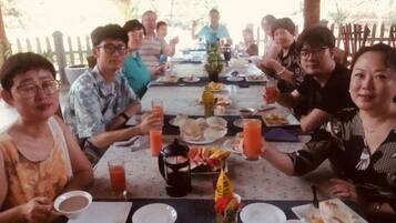 Un grupo de personas sentadas alrededor de una mesa con comida y bebidas, bajo un techo de paja.