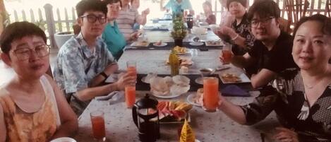 A group of people seated around a table with food and drinks, under a thatched roof.