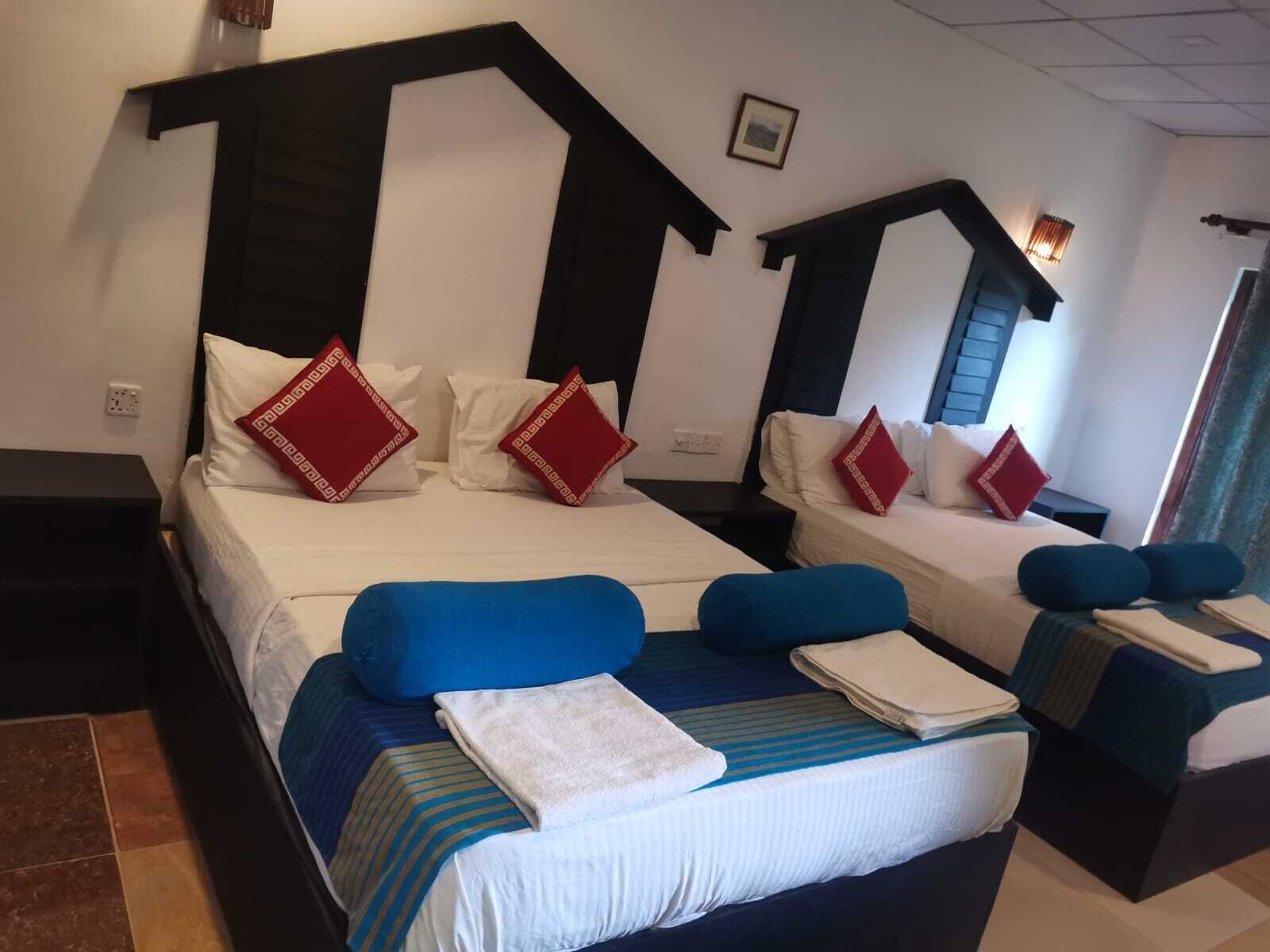 A hotel room with two beds, each with a distinct headboard design. The room has a tiled floor and a framed picture on the wall.