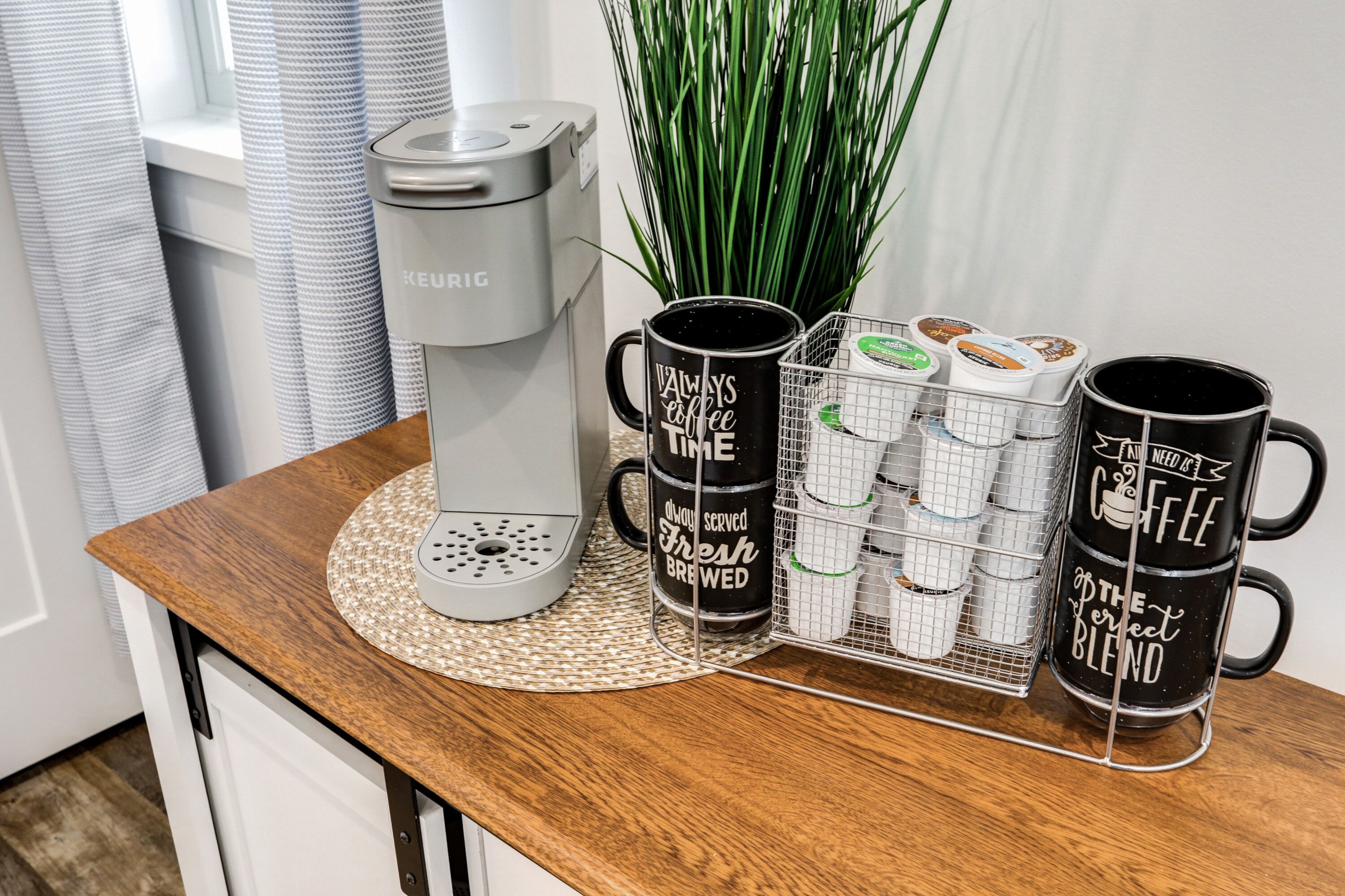 Keurig-kahvinkeitin puupintaisella työtasolla, kaksi mustaa kahvimukia, joissa on tekstiä, ja metallinen kori, jossa on kahvikapseleita.