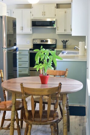 A kitchen with white cabinets, a stainless steel refrigerator, a microwave, and an oven. There is a wooden dining table with chairs and a potted plant on it.