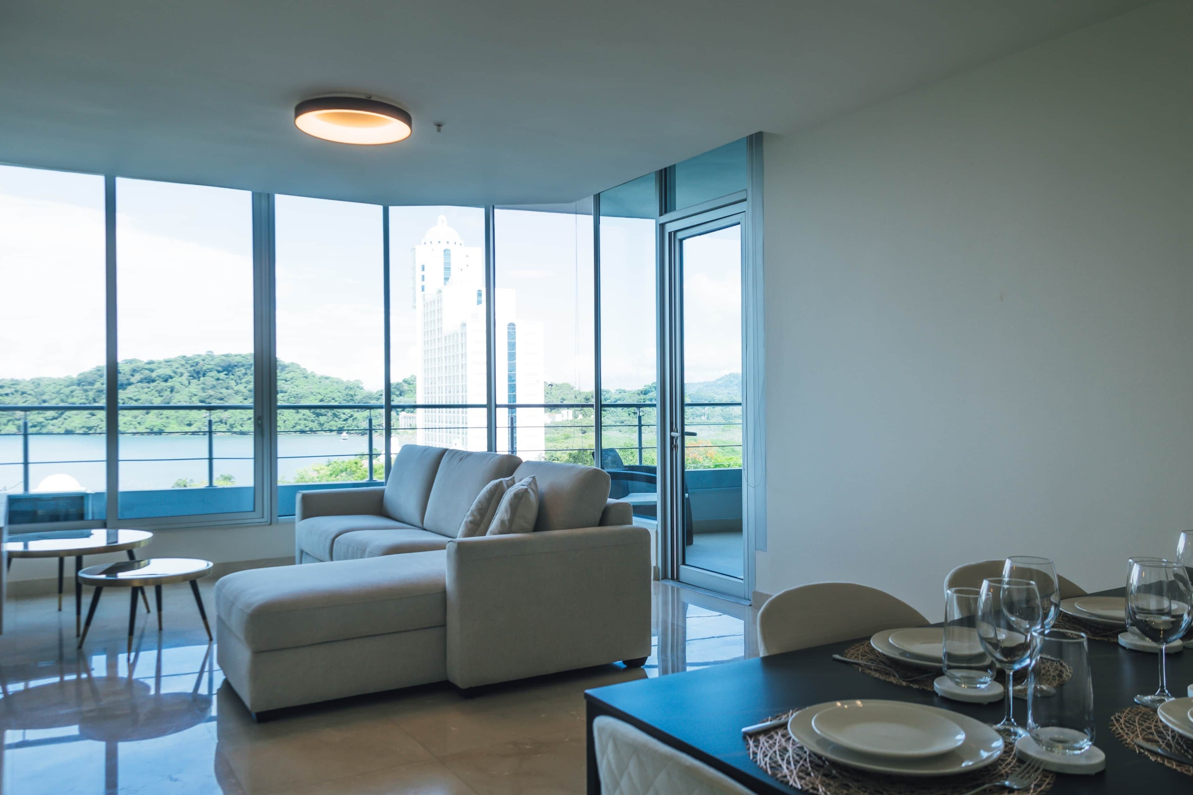 Une chambre moderne dotée d’un coin repas, d’un canapé et de grandes fenêtres donnant sur un immeuble et des espaces verts.