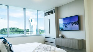 A modern hotel room with a large bed, a TV mounted on the wall, and a view of a cityscape through large windows. - Apartment in Casa Bonita (Veracruz)