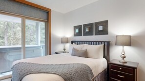 A bedroom with a large bed, two bedside tables with lamps, a window with a view of trees, and three wall-mounted picture frames. - Kookaburra #304 | 3 Peaks Rentals (Sun Peaks)