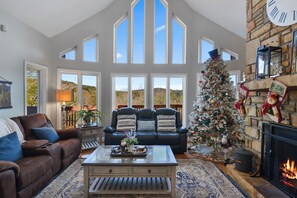 A spacious living room with a fireplace, a Christmas tree, and a large window overlooking a scenic landscape.