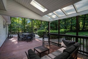 A covered outdoor deck with a table and chairs, surrounded by greenery.