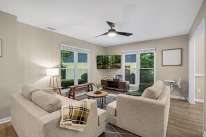 A living room with a ceiling fan, a sofa, a coffee table, a TV, and a window with a view of greenery.