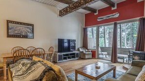 Ein gemütliches Wohnzimmer mit einem großen Fernseher, bequemen Sitzgelegenheiten und Blick ins Freie durch große Fenster.
