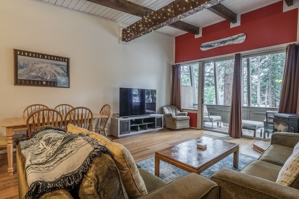 A cozy living room with a large TV, comfortable seating, and a view of the outdoors through large windows.