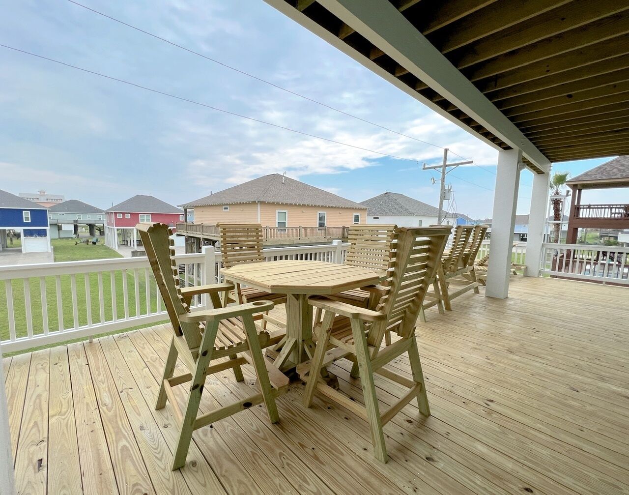Une terrasse en bois avec une table ronde et des chaises, un espace couvert avec une rambarde, et des maisons en arrière-plan.