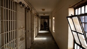 Um corredor comprido e estreito, com portas gradeadas de um lado e uma janela do outro.