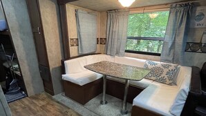 Interior compacto de uma motorhome com mesa de jantar, assentos acolchoados, janela com cortinas e iluminação no teto.